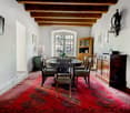 Dining room with large round table, wooden ceiling, white walls, wooden cabinets, floor lamp, large red carpet, windows