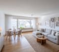 modern living room with wooden floor, large windows, round dining table, gray sofa, coffee table, lamps, rug, framed pictures on the wall, potted plants