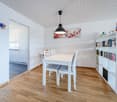 Dining room with white table and chairs, wooden floor, white walls, white ceiling, white bookshelf, white kitchen cabinets, hanging lamp, picture frame, gnomes on bookshelf