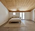 Bedroom: a wooden bed with white linens, two nightstands with lamps, white walls, wooden ceiling, tiled floor, a carpet, two windows, and a glass door leading to a balcony.
