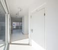 Empty hallway with white tiles, glass doors, and a balcony visible in the distance.