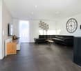 Contemporary living room with black leather sofa, wooden console table, flatscreen television, large clock, white curtains, wooden stools, wooden floor, black wall and white walls.