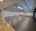 Empty room with white walls, gray floor, white ceiling with beams, large windows, and a door.