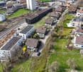 Aerial view of a residential area with several houses and buildings, some with solar panels on the roofs, surrounded by green grass and trees, with a road and a railway track in the area.
