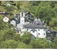 A picturesque village nestled in a lush, green valley. The buildings are made of stone and have traditional alpine architecture, with a church tower and several multi-story structures. The village appears to be built on a hillside, with some buildings hav