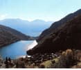 The image shows a scenic mountain landscape with a lake in the valley below. The mountains have a rugged, forested appearance, and the lake appears calm and reflective. There is a small village or town visible in the foreground, nestled among the hills. T