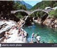 A scenic image of a stone arch bridge over a clear blue river surrounded by lush green mountains. Many people are swimming and relaxing on the rocks around the river.