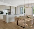 The image shows a modern, open-concept kitchen and dining area. The kitchen features light-colored cabinets, a large island with a sink, and built-in appliances including a dishwasher. The dining area has a wooden table with chairs, and the room is filled