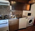 Compact kitchen with a stainless steel countertop, an electric stove, sink, and wooden cabinets. Appliances include a white microwave and a white washing machine. The wall has a tile design, and an open doorway to another room is visible on the right.