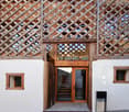 The image shows the exterior of a traditional wooden building with a lattice-style roof structure. The building has a wooden entrance door and windows, and the exterior walls are made of white plaster. The ground in front of the building is paved with cob