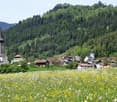 The image shows a picturesque village nestled in a lush, mountainous landscape. The village features a church with a tall spire, surrounded by traditional houses and buildings. The foreground is filled with a vibrant field of yellow and white wildflowers,