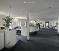 A large office space with white walls, white pillars, gray carpet, several desks, monitor, chair, glass door, white cabinets, and potted plants