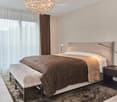 Spacious bedroom with large bed, bedside tables, lamps, chandelier, carpet, white and brown drapes, bed bench, clean white walls.