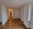 An empty hallway with wooden floors, white walls, ceiling, two sets of white wardrobes with doors, a window, and an open doorway at the end.