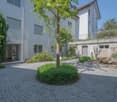 Two-story residential building with white walls and brown roof, paved courtyard with a tree in the center, plants and bushes on both sides, and a large rock with a fountain