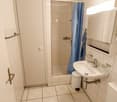 Compact bathroom with shower cubicle, white tiles, toilet, sink, mirror, towel rack, shower curtain, and small waste bin.