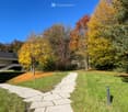 Stone pathway in the garden with a green lawn, surrounded by trees with autumnal colors, and a building in the background.