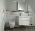 Bathroom with a white toilet, sink, white vanity with drawers, white tiled walls, wooden shelf above the toilet, mirror, radiator, and white door