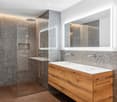 Modern bathroom with a glass shower stall, large mirror, wooden cabinet with double sinks, and gray tiled walls.