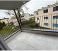 A balcony of an apartment building with tile flooring and a fence overlooking a grassy field with trees and other buildings in the distance
