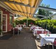 Outdoor restaurant area with yellow retractable roof, tables and chairs covered with white tablecloths, plants, red chairs, green bushes.