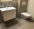 The image shows a modern bathroom with a white vanity sink, a toilet, and gray tile flooring. The bathroom has a clean and minimalist design with chrome fixtures and accessories.