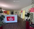 Wooden floor, modern design desk, red potted plants, entrance signage with logo, bright lighting, minimalist decor, open hallway, yellow walls, glass doors, minimalistic art