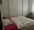Bedroom with a double bed, white walls, white bed frame, red bed sheets, floral bedspread, white pillow, white nightstand, white lamp, white sliding wardrobes, wooden floor, white ceiling lamp, small wooden table, green cloth, chair, white curtains
