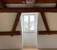 An attic with wooden beams, a white wall, a carpeted floor, and a window with an outdoor view.