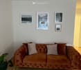 a living room with a brown couch, wooden floor, white wall, a rug, plant, picture frames