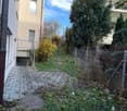 Brick pathway leading to a garage with a concrete ceiling, surrounded by shrubs, and a small garden with plants and trees.