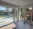 Modern living space with large glass walls, overlooking a garden with greenery and a wooden shed, tiled floor.