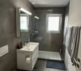 Gray walls, white tiles, glass shower stall, white porcelain toilet, white vanity with sink, a roll of tissue paper, a towel rack with towels, a radiator, blue bath mat.