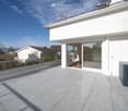 A modern white building with an open terrace, equipped with glass doors and railings, overlooking a sunny landscape with other houses.