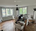 A wooden floor, white walls, desk, two monitors, books, stack of books, chairs, radiators, lamps, and two windows