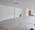 Empty room with grey tiled floor, air conditioning vent on the ceiling, white walls, electrical outlet, and an entrance in the background.