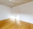 A square shaped room with wooden floors, white walls, an open door to another room with a white and gray floor, and lights on the ceiling