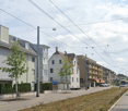 Urban street scene with residential buildings, tram tracks, trees, and a tram. Multistory residential building on the left, other residential buildings and tram tracks in the background.