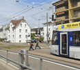 urban street with tram, buildings, traffic signals, people walking, median with guard rails, overhead wires, and parked cars