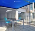 Covered outdoor patio with blue retractable awning, glass railing, and modern patio furniture including a table and blue chairs.