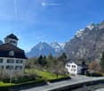 Swiss house with mountains in the background, large windows, brown roof, small garden, driveway
