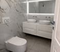modern bathroom with white marble tiles, two wash basins, a toilet, and a mirror.