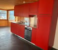 modern kitchen with red cabinets, integrated dishwasher, oven, kitchen island with sink and faucet, stainless steel countertops, and wooden wall paneling
