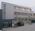 modern building, grey and white facade, large windows, glass doors, paved parking area, two cars parked