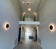 Modern hallway with grey tiled floor, white walls, recessed lighting, and a suspended light fixture with multiple circular bulbs. Features a mirrored balcony overlook and built-in wall-mounted lighting on both sides.