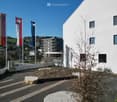 White building with glass windows, flags, paved area, small garden, another building in the background