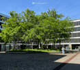 Modern multi-story building with large windows and a landscaped courtyard featuring trees and shrubs.