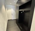 Modern hallway with dark grey tiled floor, white walls, built-in black storage unit with shelves and drawers, and a white door at the end.