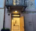 The front entrance of a residential building with a wooden door. It features a balcony above the door, with wrought iron railings and an ornate design. The number '6' is visible on a plaque above the door, and a lamp illuminates the entrance. The interior
