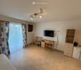 A furnished room with a bed, desk, chairs, television, floor lamp, and cabinet. The room has tiled flooring and a window with curtains.